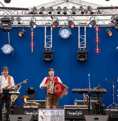 Eine Band spielt auf einer Bühne mit blauer Rückwand. Die Musiker tragen traditionelle Kleidung und spielen verschiedene Instrumente, während bunte Lichter leuchten.