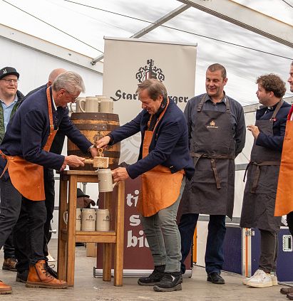 Eine Gruppe von Menschen in traditioneller Kleidung zapft ein Fass Bier. Hinter ihnen steht ein Banner mit einem Wappen. Einige tragen orangefarbene Schürzen.