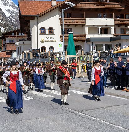 Eine traditionelle Blaskapelle in Trachten marschiert bei sonnigem Wetter durch eine Straßenkulisse in einem Alpenort, während Menschen am Straßenrand ihnen zuschauen.