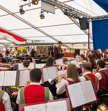 Eine Blaskapelle in traditionellen Trachten spielt in einem großen Festzelt. Die Musiker sind mit Notenblättern beschäftigt, während Zuschauer im Hintergrund stehen.