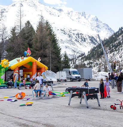 Eine Winterlandschaft in den Bergen mit schneebedeckten Gipfeln. Eine Gruppe von Menschen genießt bunte Hüpfburgen und Spiele im Freien auf einem Platz.