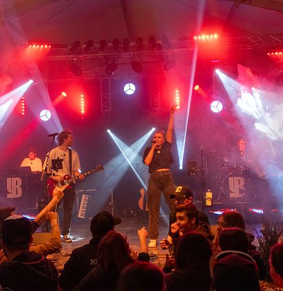 Eine Band tritt bei einem Konzert auf der Bühne auf, umgeben von bunten Lichtern und Nebel. Die Musiker spielen leidenschaftlich und das Publikum genießt die dynamische Atmosphäre.