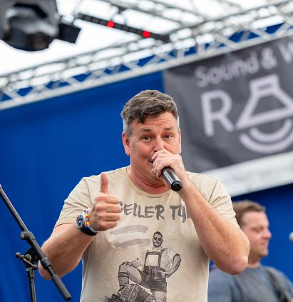 Ein Mann steht auf einer Bühne und singt in ein Mikrofon. Er trägt ein T-Shirt mit einem Aufdruck und zeigt mit der Hand ein Daumen-hoch-Zeichen. Im Hintergrund ist ein blauer Vorhang.