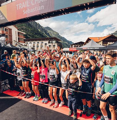Eine Gruppe von Kindern stehen unter einem Startbogen für den PKids beim Pitz Alpine Glacier Trail im Freien. Die Teilnehmer heben die Arme, um zu jubeln. Gebäude und Berge sind im Hintergrund.