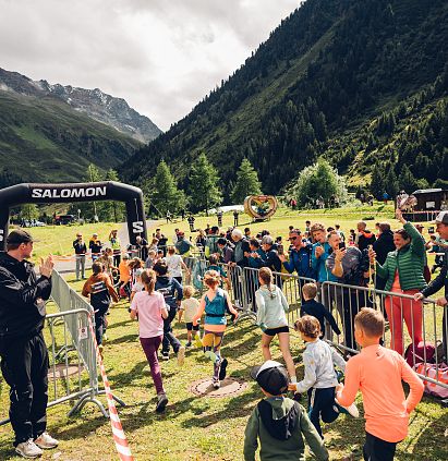 Gruppe von Kindern laufen durch einen Parcour in bergiger Landschaft, umgeben von Zuschauern und Bäumen.
