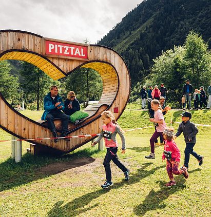 Mehrere Kinder laufen und auf einer grünen Wiese neben einem herzförmigen Holzrahmen mit der Aufschrift &quot;Pitztal&quot; in den Bergen.