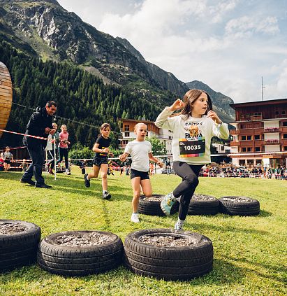 Kinder nehmen am PKids-Lauf des Pitz Alpine Glacier Trail teil. Sie springen über Autoreifen auf einer grünen Wiese, umgeben von Bergen und jubelnden Zuschauern.