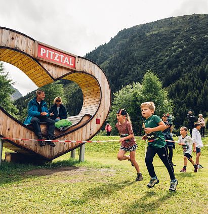 Kinder laufen fröhlich beim PKids beim Pitz Alpine Glacier Trail. Im Hintergrund sitzen Personen in einem herzförmigen Holzrahmen mit der Aufschrift Pitzal.
