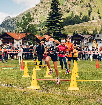 Kinder rennen beim PKids, Teil des Pitz Alpine Glacier Trails im Freien über eine Wiese mit Hindernissen. Umgeben von Zuschauern vor einer Berglandschaft.