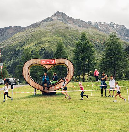 Kinder laufen auf einer Wiese vor einer herzförmigen Plattform mit &quot;Pitztal&quot;-Schild. Im Hintergrund sind Berge und Bäume zu sehen.