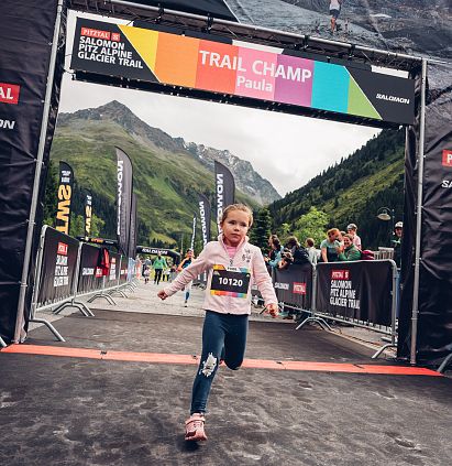 Ein Mädchen überquert die Ziellinie des PKids in einer Berglandschaft. Im Hintergrund sind Berge, Fahnen und Zuschauer zu sehen.