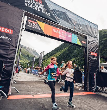 Zwei Kinder erreichen freudig den Zieleinlauf des PKids beim Pitz Alpine Glacier Trail. Im Hintergrund sind Berge und eine bunte Trail-Gangway zu sehen.