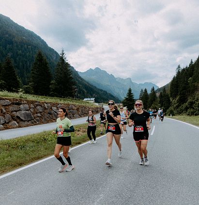 Menschen laufen auf einer Straße in einer bergigen und bewaldeten Landschaft unter bewölktem Himmel, umgeben von Natur und Bergen im Hintergrund.