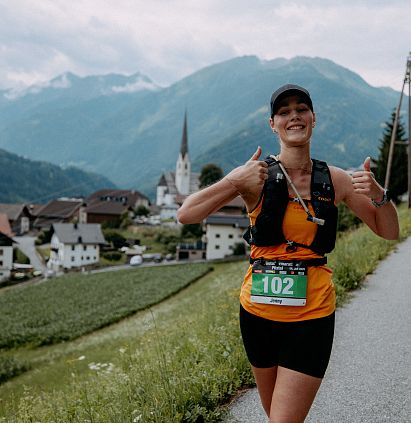 Lächelnde Läuferin mit Startnummer in sportlicher Kleidung, Daumen hoch. Berglandschaft mit Häusern und Kirche im Hintergrund. Sommerliches Wetter.