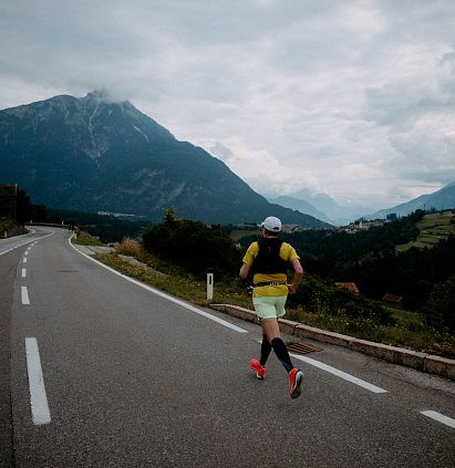 Ein Läufer auf einer leeren Landstraße, umgeben von Bergen und Wiesen. Der Himmel ist bewölkt, und die Szene wirkt ruhig und abgelegen.