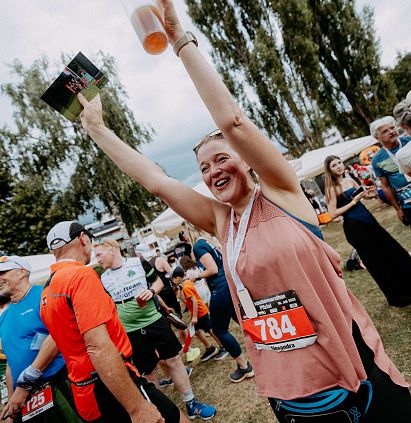 Frau jubelt bei einer Outdoor-Veranstaltung mit Gewinnermedaille und Getränk, umgeben von anderen Teilnehmern und Bäumen im Hintergrund.