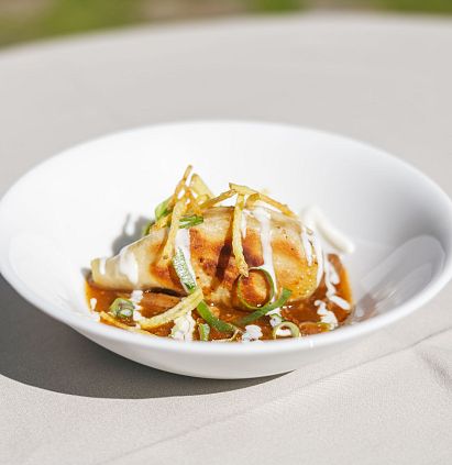 Ein Gericht auf einem weißen Teller, das eine gefüllte Teigtasche zeigt, garniert mit einer Sauce, Zwiebelstreifen und Kräutern. Der Hintergrund ist unscharf.