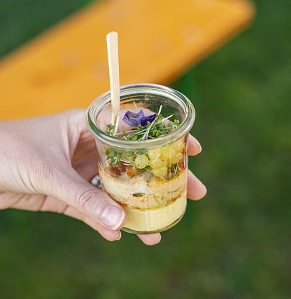 Eine Person hält ein Glas mit schichtigem Essen in der Hand. Das Glas enthält eine bunte Mischung aus frischem Gemüse, Kräutern und einer cremigen Schicht.