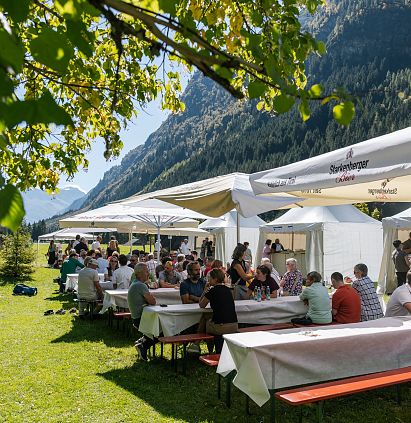 Eine sommerliche Szene mit vielen Menschen, die an langen Tischen unter Zelten im Freien sitzen. Umgeben von Grünen Wiesen und Bergen, genießen sie wahrscheinlich ein Fest oder ein Event.