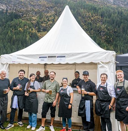 Eine Gruppe von Menschen steht vor einem weißen Zelt auf einem Festival. Sie tragen Schürzen und halten Getränke, umgeben von einer malerischen Berglandschaft.