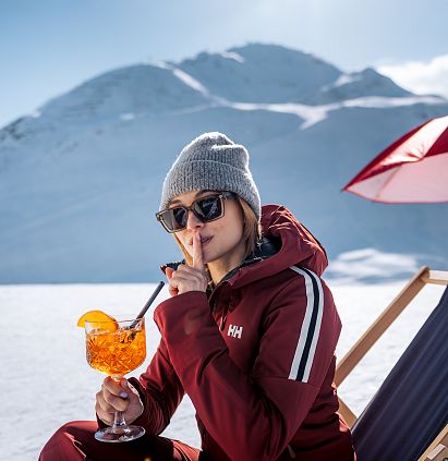 Eine Person in Winterkleidung sitzt auf einem Liegestuhl im Schnee. Sie genießt einen Cocktail und hat den Finger vor den Lippen. Im Hintergrund sind schneebedeckte Berge.