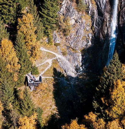 Luftaufnahme eines Wasserfalls in einem bewaldeten Gebirgsgebiet im Herbst. Ein kleiner Weg schlängelt sich durch die Bäume, die in leuchtenden Gelb- und Orangetönen stehen.