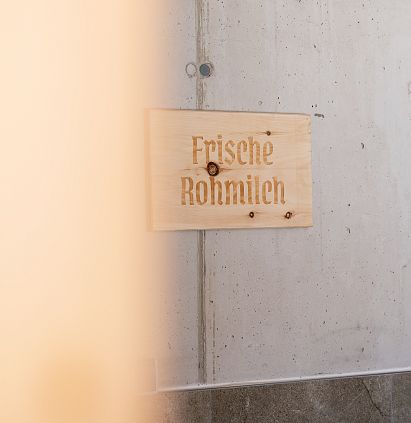 A wooden sign bearing the words ‘Fresh Raw Milk’ attached to a concrete wall. The foreground is out of focus, which gives the picture an interesting sense of depth.
