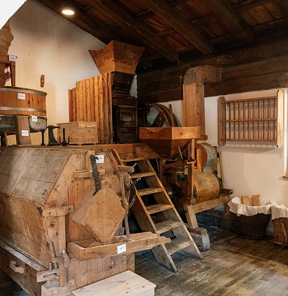 A rustic room with traditional wooden machinery, a wooden granary and old tools on wooden shelves. Daylight streams through a window onto the wooden floor.