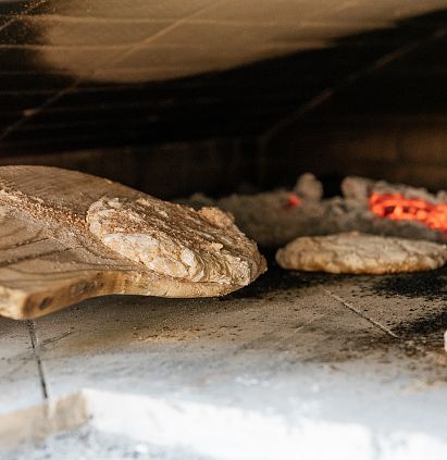 A wood-fired oven with glowing embers, in which several round, unbaked loaves of bread dough are resting on a wooden peel. The loaves are baked deep inside the oven, surrounded by heat.