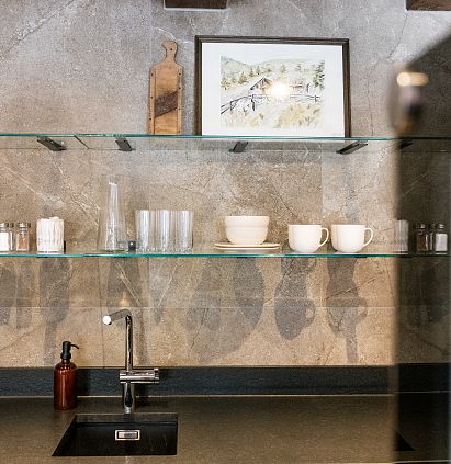 A modern kitchen area with a grey stone wall, glass shelves displaying cups and glasses, a black sink and an ornate picture on the wall.