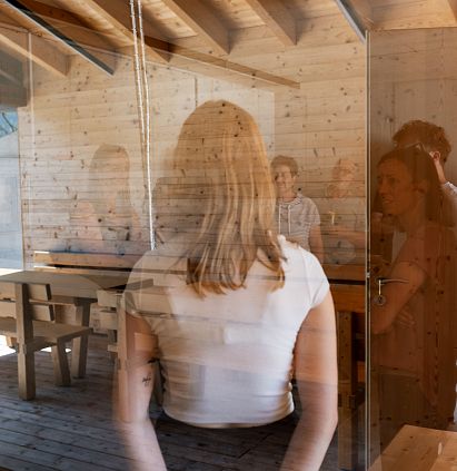 A woman with long hair is standing with her back to the camera in a wood-panelled room, which is separated by a glass wall. Inside, other people are sitting at a table.