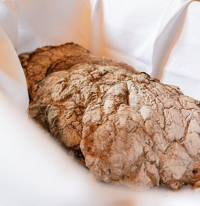 Crispy, light-brown bread in a white bread basket, resting on a napkin. The crust is uneven and cracked, suggesting a rustic, artisanal production.