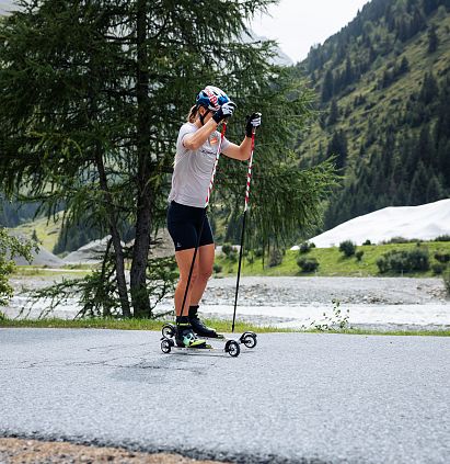 A person wearing a helmet and carrying poles is roller-skying along a road in an alpine landscape. In the background, trees and a white hill, which is a snow storage area, can be seen.