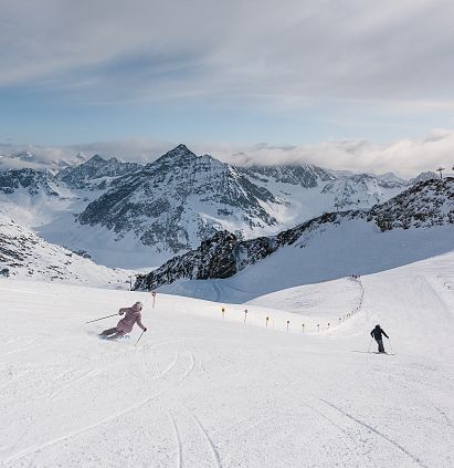 Zwei Personen fahren Ski auf einer schneebedeckten Piste in den Alpen. Im Hintergrund sind majestätische Berge und ein teils bewölkter Himmel zu sehen. Die Atmosphäre ist klar und kalt.