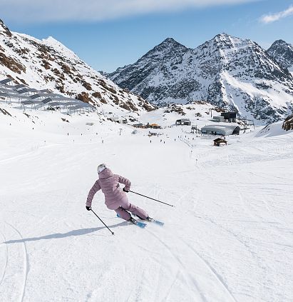 Eine Person in rosa Skibekleidung fährt schwungvoll eine breite Skipiste hinunter. Umgeben von schneebedeckten Bergen erstreckt sich der blaue Himmel über der Winterlandschaft.