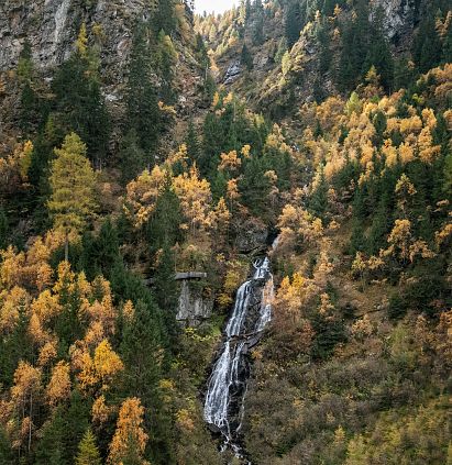 Ein Wasserfall fließt durch einen bewaldeten Berghang im Herbst. Die Bäume leuchten in Gelb- und Grüntönen, während das Wasser zwischen Felsen hinabstürzt.