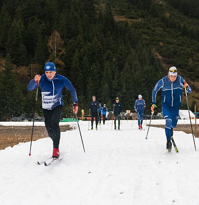 Zwei Langläufer in blauen Anzügen und Helmen gleiten dynamisch über eine schneebedeckte Strecke. Im Hintergrund sind Bäume und mehrere Menschen erkennbar.