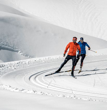 Zwei Langläufer, einer in einer orangefarbenen Jacke, der andere in einer blauen Jacke, gleiten an einem hellen, klaren Tag über präparierte Schneeloipen.