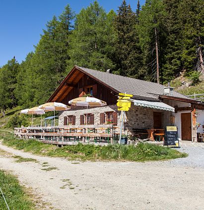 Eine rustikale Berghütte mit einer Holzveranda und Sonnenschirmen. Umgeben von grünen Wäldern und einem schmalen Weg, der zur Hütte führt.