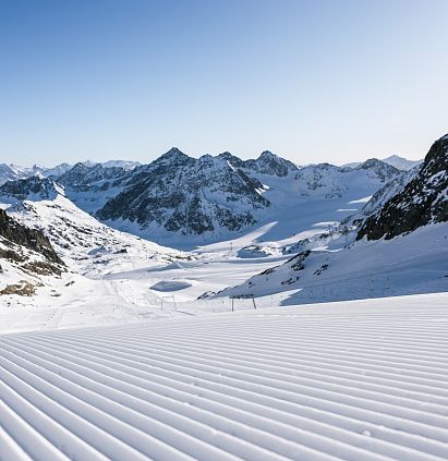 Verschneite Berglandschaft unter klarem Himmel. Im Vordergrund sind parallele Linien auf frisch präpariertem Schnee zu sehen. Die Berge erstrecken sich bis zum Horizont.