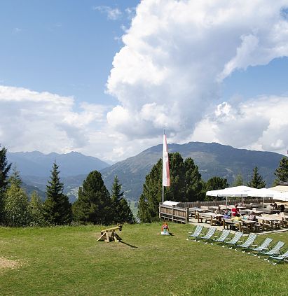 Eine idyllische Berglandschaft mit einem Holzhaus im Vordergrund. Auf der Wiese stehen Liegestühle, ein Spielplatz mit Schaukel und im Hintergrund erstrecken sich Berge und ein blauer Himmel.