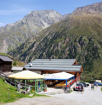 Eine alpine Landschaft mit einem Holzgebäude, einem Gelben Sonnenschirm und mehreren Besuchern. Im Hintergrund erheben sich hohe Berge unter einem blauen Himmel.