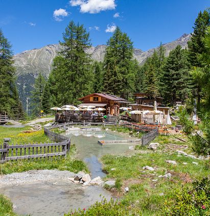 Ein malerisches Bergrestaurant in den Alpen, umgeben von hohen Pinien und grünem Gras. Sonnige Terrasse mit Tischen, Berglandschaft im Hintergrund, blauer Himmel.