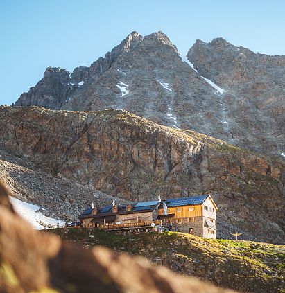 Eine malerische Berghütte auf einem steilen Hang, umgeben von majestätischen Bergen. Der Himmel ist blau und die Sonne scheint, was die Landschaft beleuchtet.