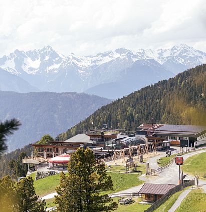 Eine Berglandschaft mit verschneiten Gipfeln im Hintergrund. Im Vordergrund sieht man ein modernes Gebäude, umgeben von Bäumen und Wiesen, ideal für Freizeitaktivitäten.