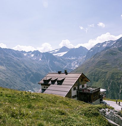 Eine malerische Berglandschaft mit einem modernen, rustikalen Gebäude im Vordergrund. Im Hintergrund erheben sich hohe, schneebedeckte Berge unter einem blauen Himmel.