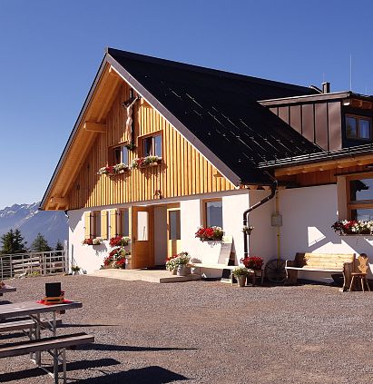 Das Bild zeigt ein modernes, rustikales Gebäude in Berglandschaft. Es hat ein schräges Dach, große Fenster und Blumen am Fenster. Tische sind im Freien aufgestellt.
