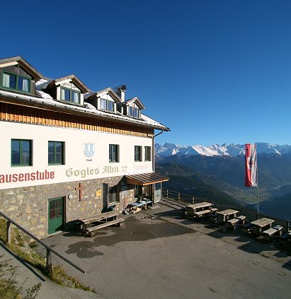 Eine Berglandschaft mit einem rustikalen Gasthaus im Vordergrund. Das Gebäude hat ein traditionelles Dach und einige Tische im Freien. Im Hintergrund sind schneebedeckte Berge zu sehen.