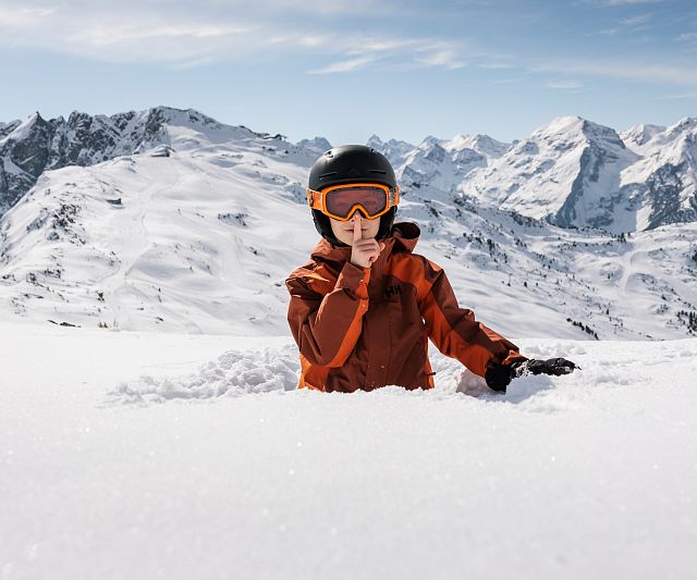 Eine Person in oranger Skijacke und Helm steht knietief im Schnee auf einem verschneiten Berg, umgeben von einer beeindruckenden Alpenlandschaft unter blauem Himmel.