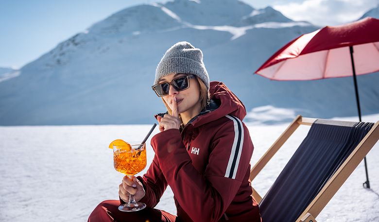 Eine Person in Winterkleidung sitzt auf einem Liegestuhl im Schnee. Sie genießt einen Cocktail und hat den Finger vor den Lippen. Im Hintergrund sind schneebedeckte Berge.